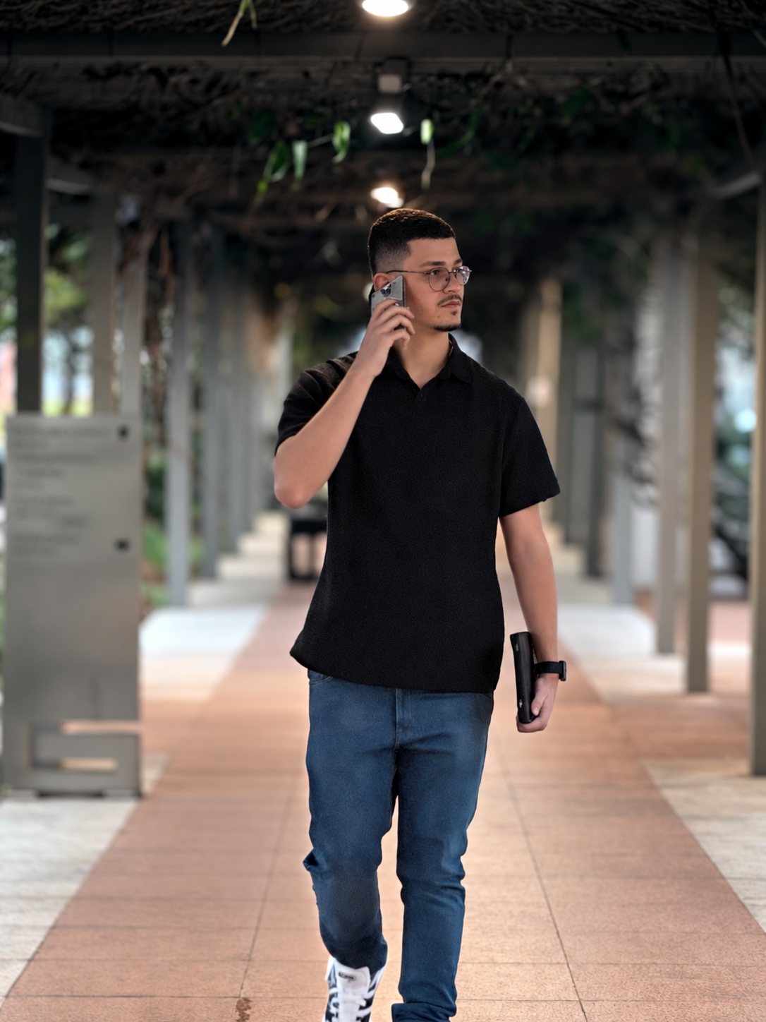 Abdeel caminhando por um corredor coberto, de óculos e camisa preta, falando ao celular